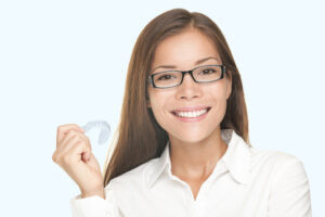 Woman in glasses holding invisible teeth aligners and smiling.